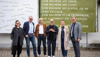 Maíra Wiener (Projektmitarbeiterin Matchbox), Werner Baumann (Ortsbürgermeister Maxdorf), Paul Platz (Kulturleiter Rhein-Pfalz-Kreis), Robert Montoto (Leiter Kulturbüro der Metropolregion Rhein-Neckar GmbH), Lea Gerschwitz (Projektleitung Matchbox), Clemens Körner (Landrat Rhein-Pfalz-Kreis) vor von Mia & Eric gestalteten Plakaten in Maxdorf | © © Arthur Bauer sechs Personen posieren für ein Gruppenfoto vor grün-weißen Plakaten mit Schrift der Künstler:innen | © © Arthur Bauer