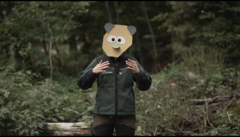 Videostill aus "In a Strange Place" | © Mia & Eric Eine Person steht im Wald und gestikuliert, sie trägt eine bunte Maske aus Pappe, die ein Fabelwesen darstellt. | © Mia & Eric