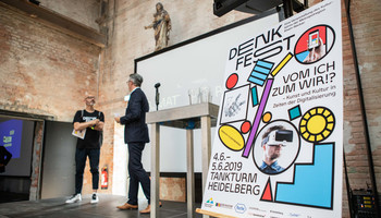 Denkfest Eröffnung 2019 im Tankturm und Betriebswerk Heidelberg, Robert Montoto und Matthias Rauch | © Arthur Bauer Im Vordergrund ein Plakat zur Bewerbung des Denkfests, im Hintergrund zwei Personen auf einer Bühne | © Arthur Bauer