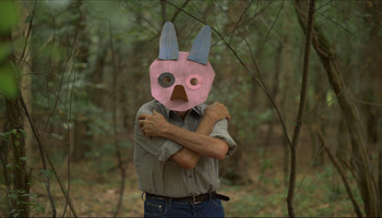 Videostandbild aus den Filmaufnahmen im Maxdorfer Heidewald im Rahmen von 3 WOODS | © Mia & Eric Ein älterer Mann steht im Wald, die Arme vor der Brust überkreuzt, er trägt eine Maske aus Pappe, die einen abstrakten Schweinekopf darstellt. | © Mia & Eric