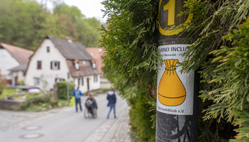 Hintergrund: Straße mit mehreren Personen, davon einer im Rollstuhl Vordergrund: Aufkleber des Camonio Inclusio Wanderwegs an einer Straßenlaterne mit Büschen dahinter. | © VRRN/ Christian Buck